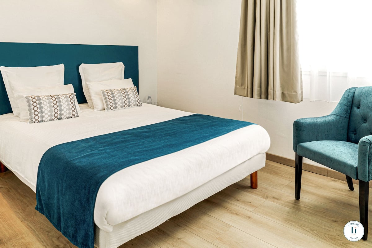 Una cama bien hecha con un detalle en color verde azulado, una silla azul y un suelo de madera.
