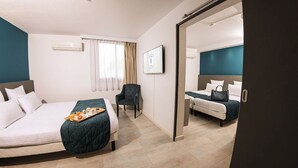 Habitación de hotel con una cama grande, una cama más pequeña, una silla, un escritorio y un baño visible a través de una puerta abierta.