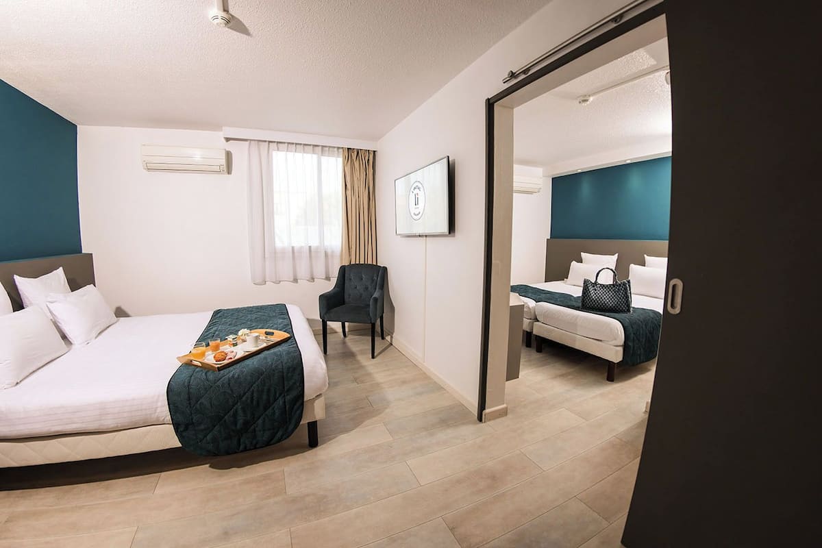 Habitación de hotel con una cama grande, una cama más pequeña, una silla, un escritorio y un baño visible a través de una puerta abierta.