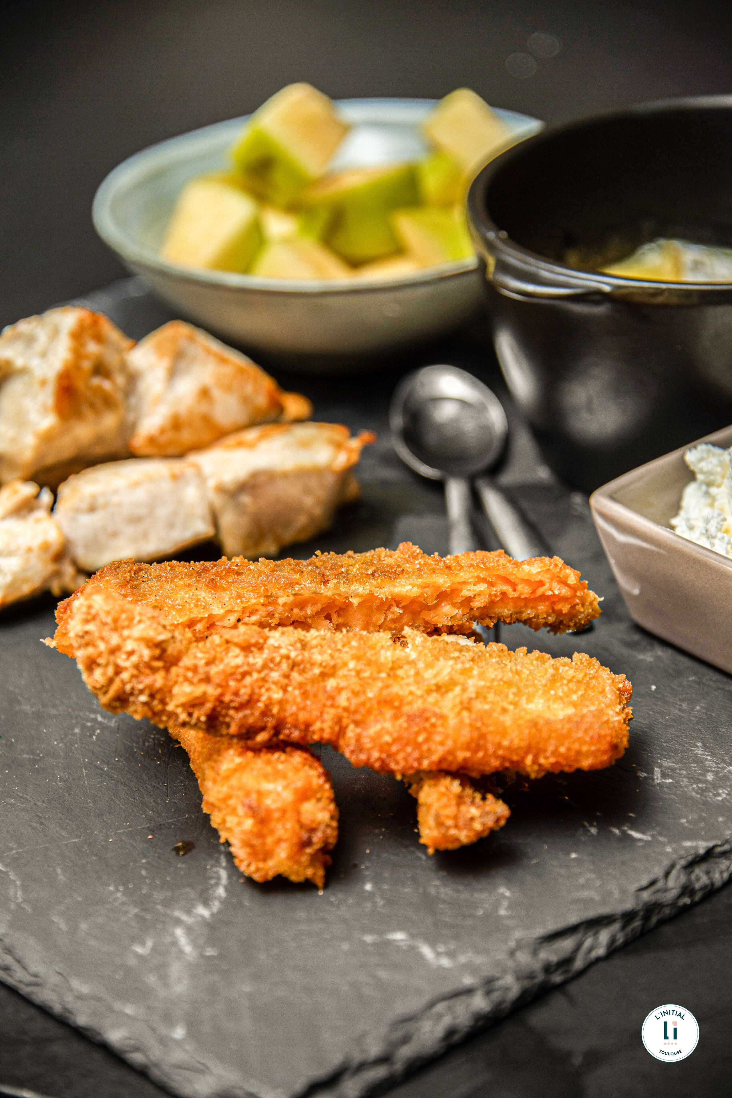 A slate serving board with breaded chicken tenders, a bowl of fruit, and a bowl of dip.