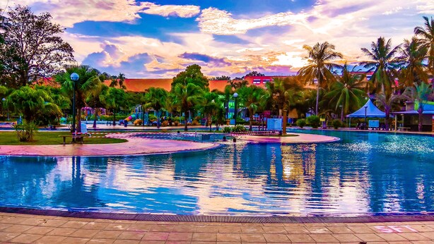 A swimming pool with a tiled deck, surrounded by palm trees and a sunset in the background. - De Rhu Beach Resort (Kuantan)