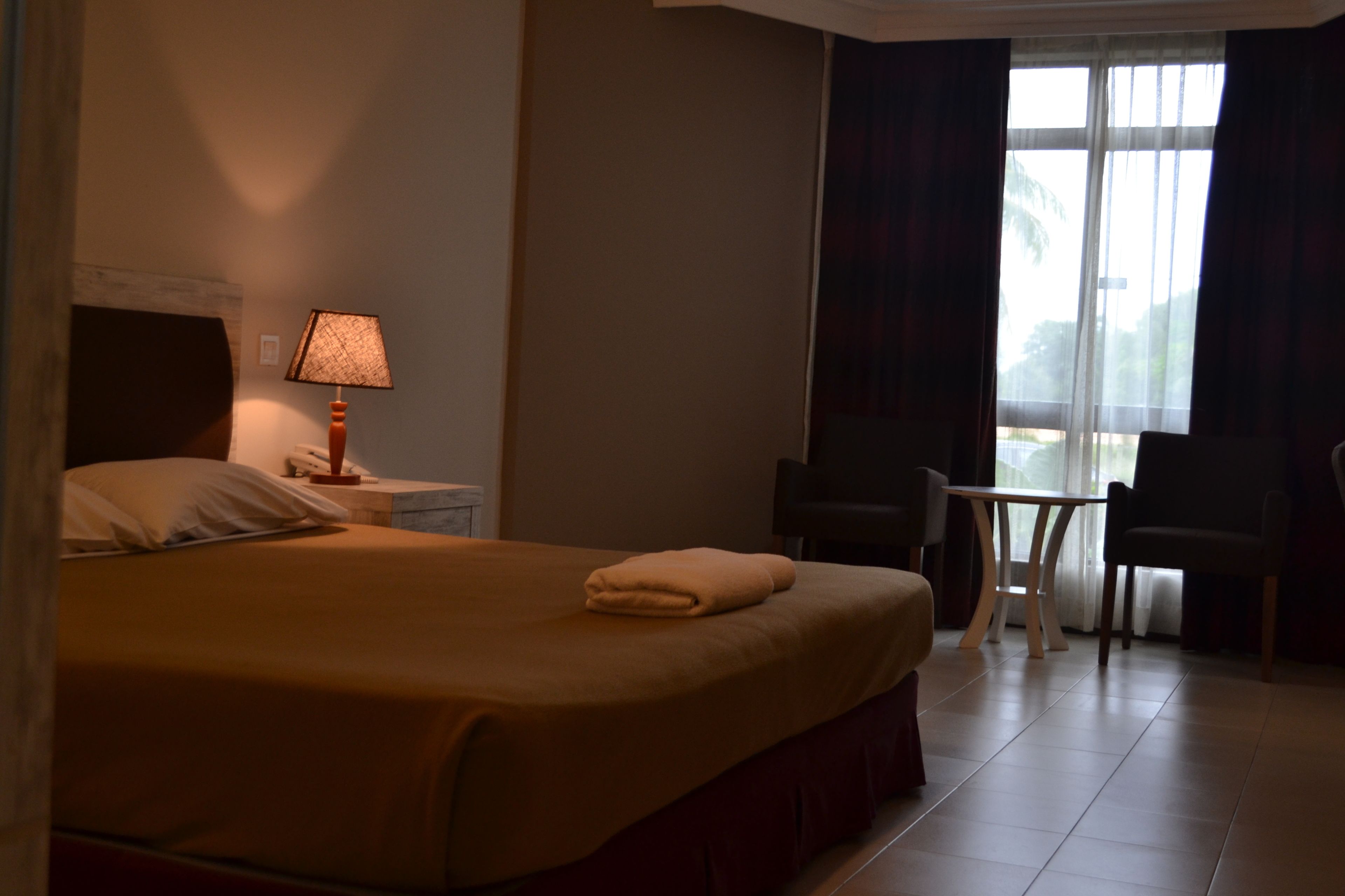 A hotel room with a bed, a nightstand with a lamp, a small table, two chairs, and a window with curtains.