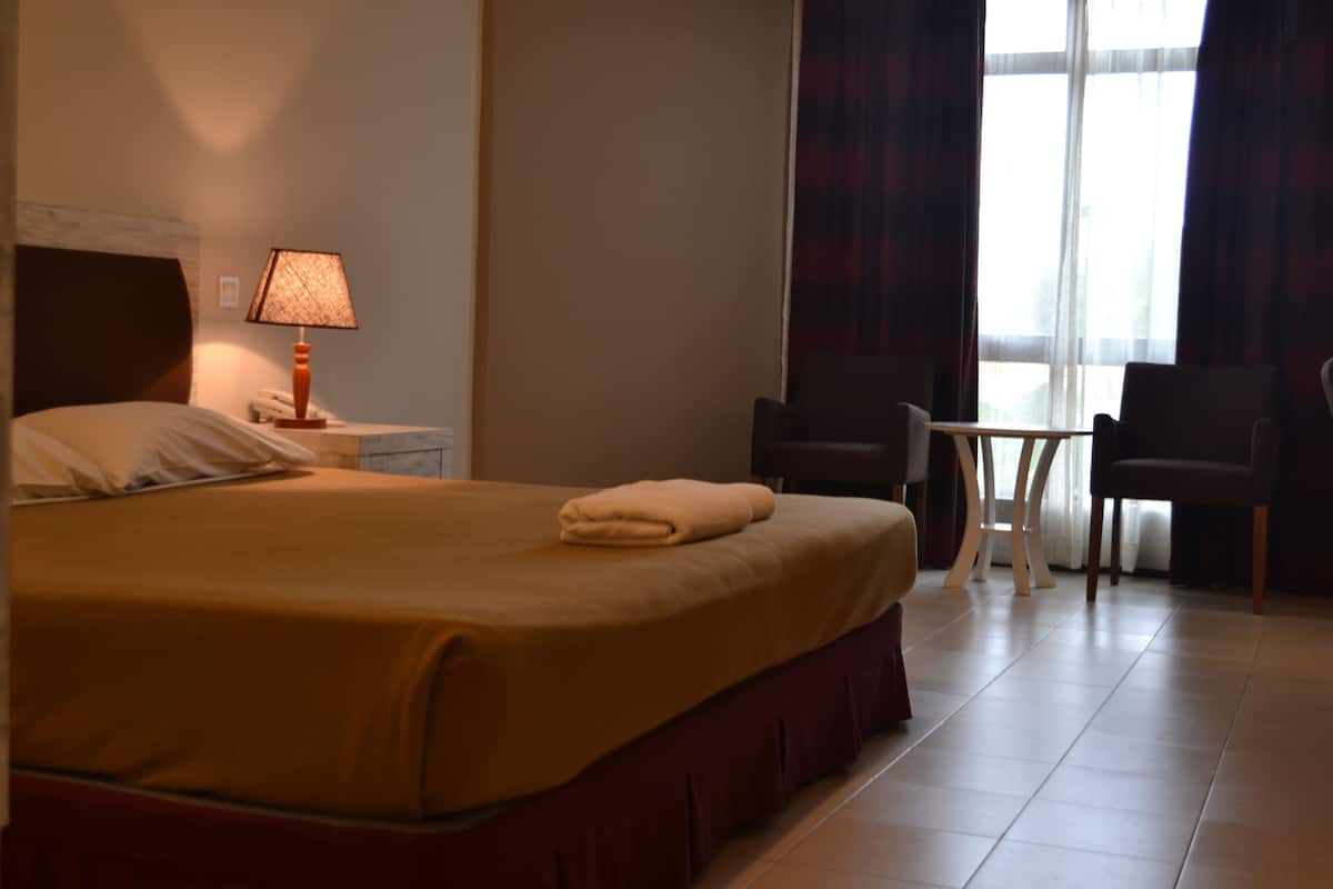 A hotel room with a bed, a bedside lamp, a small table, two chairs, and a window with curtains.