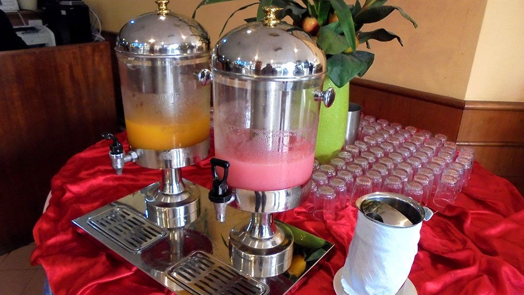 A beverage station with two dispensers, one with a yellow drink and another with a pink drink, and a tray with a metal cup.