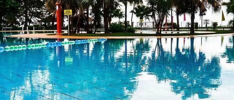 A swimming pool with clear blue water, surrounded by palm trees and a tiled deck.