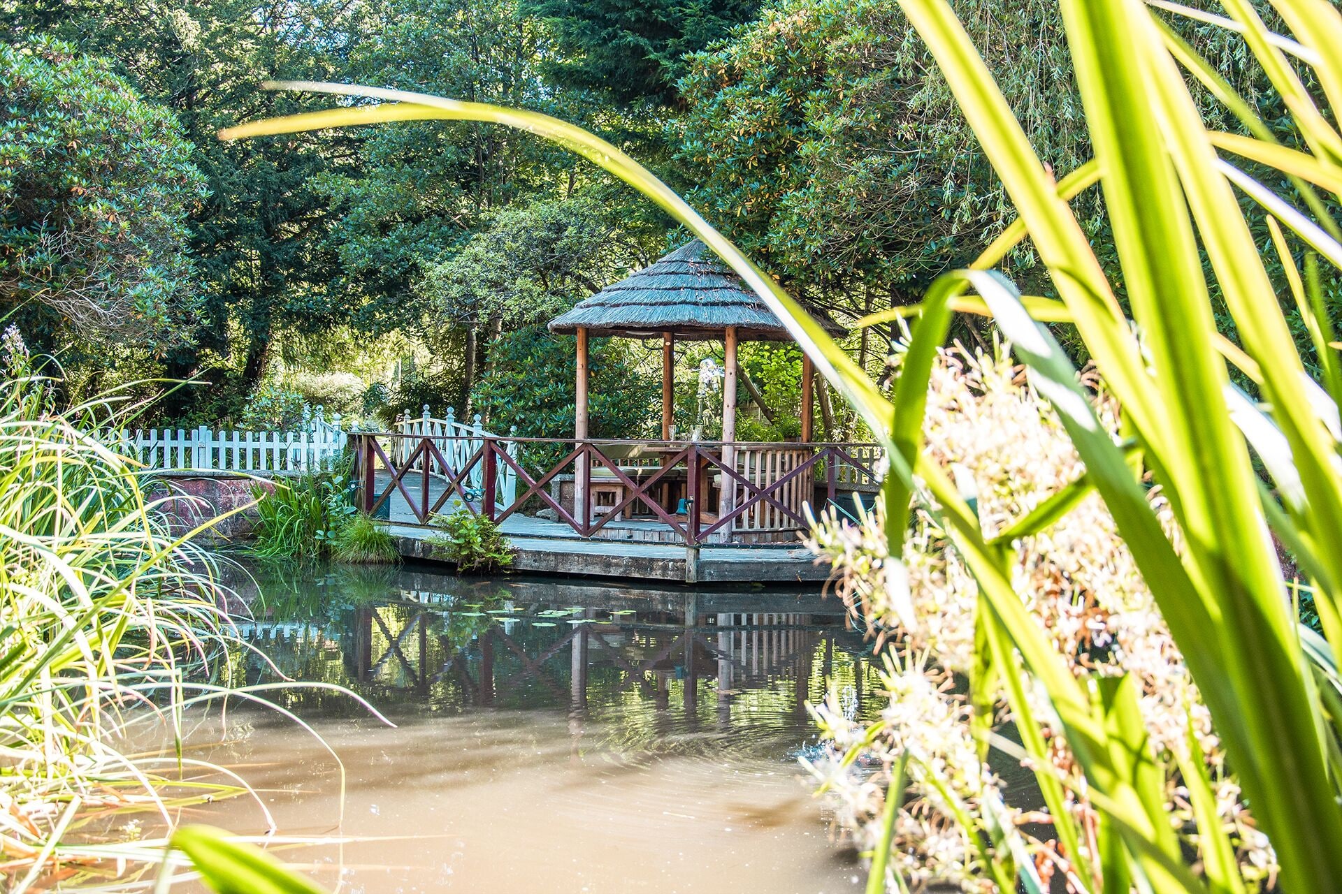 A gazebo with a thatched roof, surrounded by a wooden fence and a small pond.