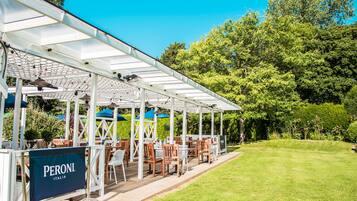 Un patio exterior con mesas y sillas, una pérgola con techo de cristal y un cartel que dice "PERONI ITALIA".