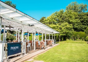 Un patio exterior con mesas y sillas, una pérgola con techo de cristal y un cartel que dice "PERONI ITALIA".