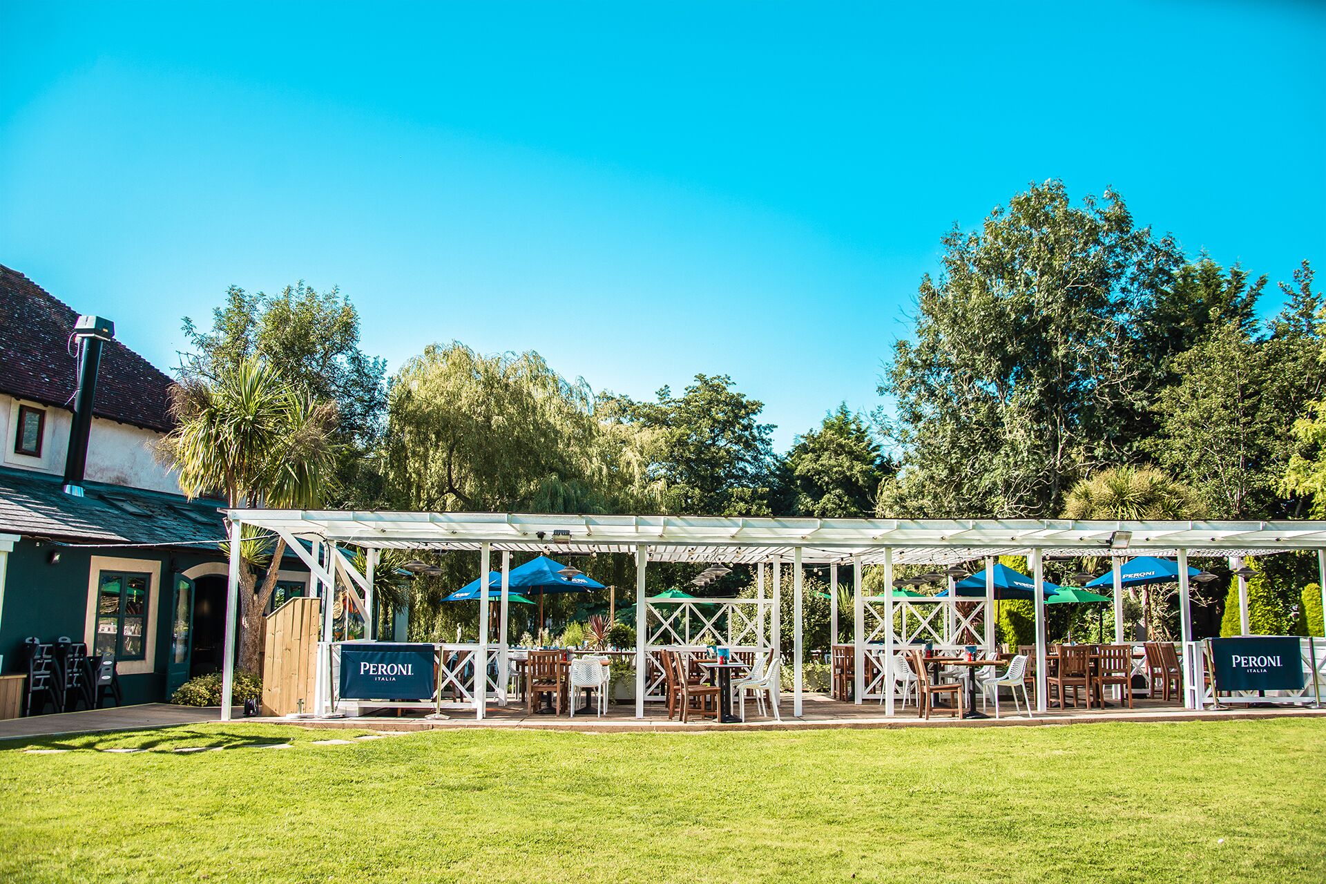 An outdoor seating area with tables and chairs under a covered structure, surrounded by greenery and trees.