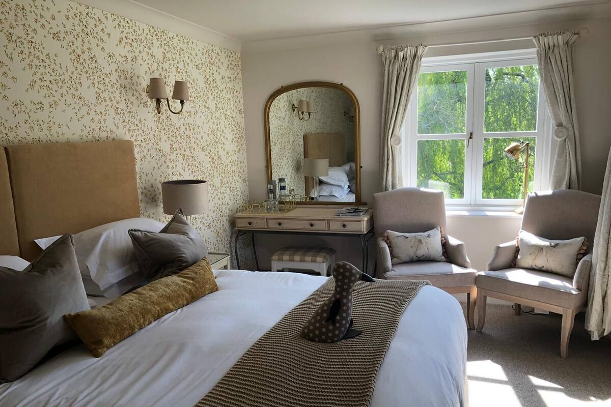 A hotel room with a large bed, a desk with a mirror, two armchairs, and a window with curtains.