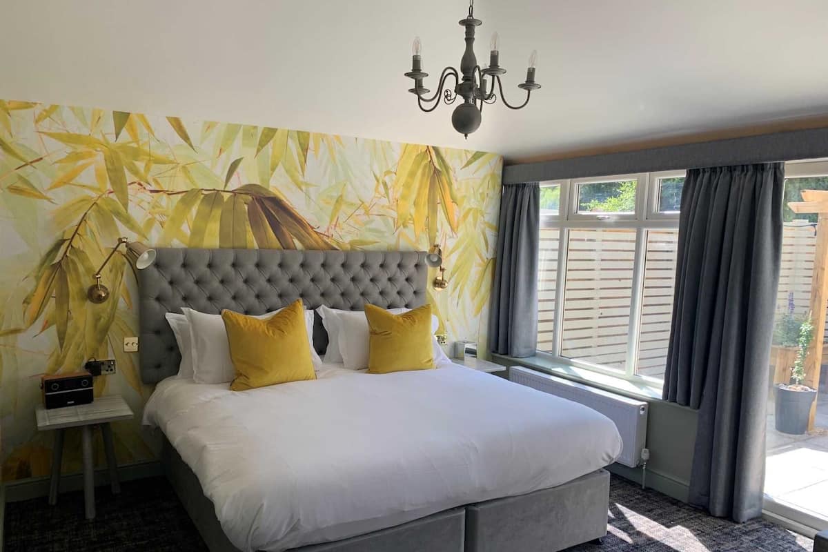 A bedroom with a tufted headboard, a large bed with white and yellow pillows, a bedside table with a lamp, and a view of the outdoors through a window with curtains.