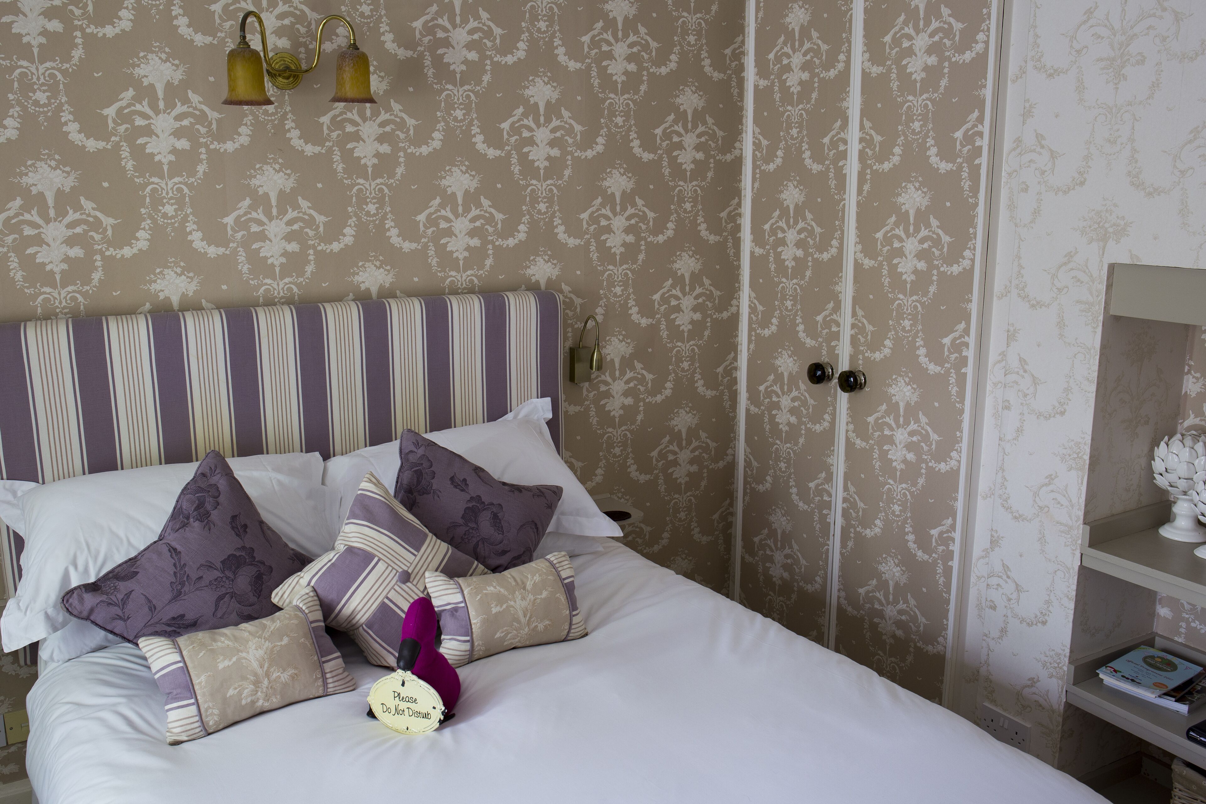 A neatly made bed with striped headboard and multiple pillows, a bedside lamp, and a small bedside table with a book and a decorative item.