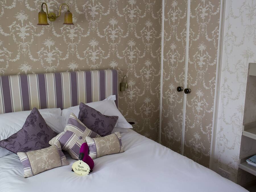 A neatly made bed with striped headboard and multiple pillows, a bedside lamp, and a small bedside table with a book and a decorative item.