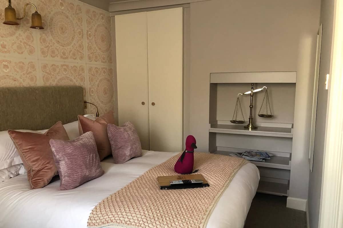 A neatly made bed with a beige blanket and pillows, a remote control, and a red decorative item on the bed. There is a wardrobe with a mirror and a shelf with a scale and lamp.