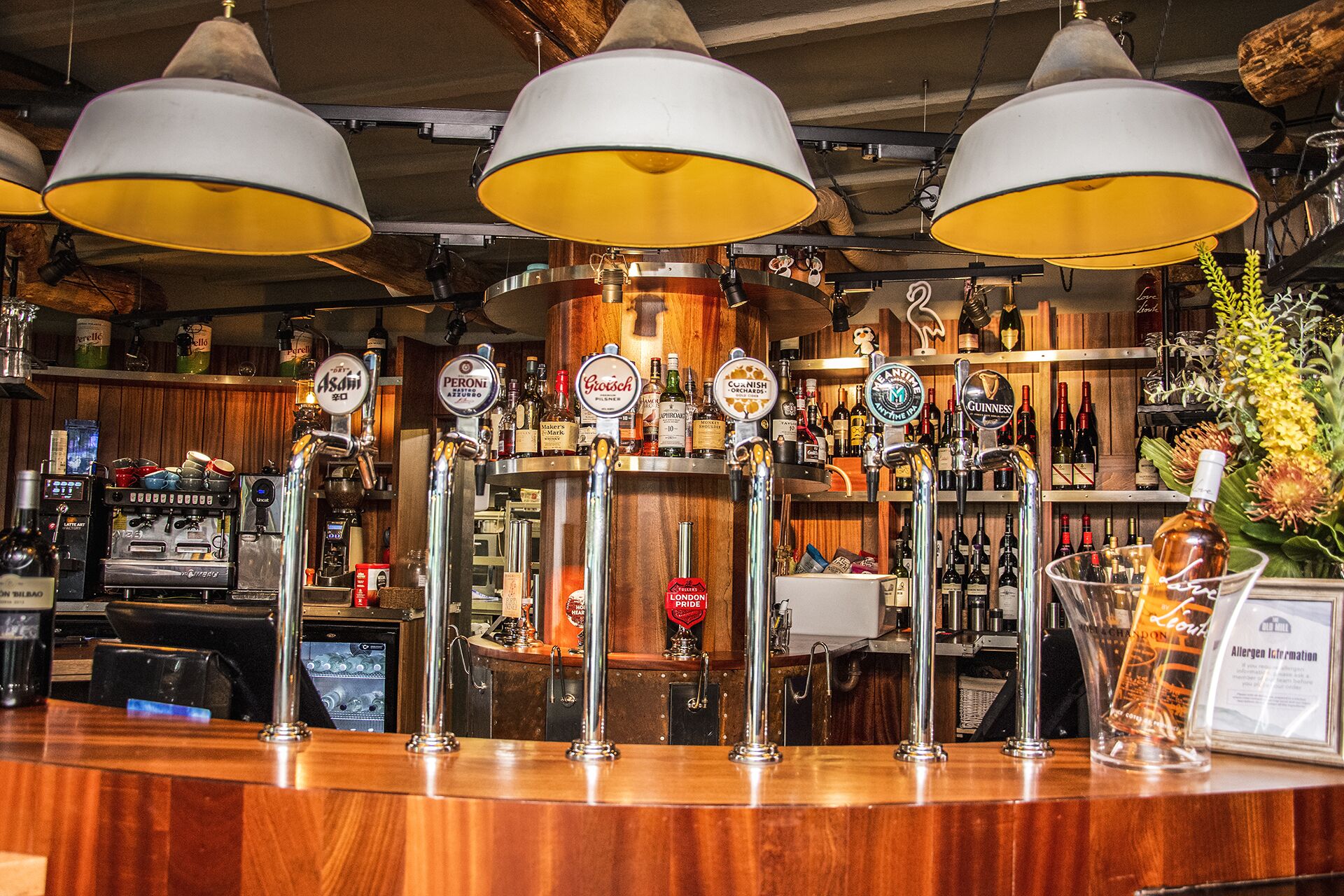 Een bar met meerdere biertaps, een koffieapparaat en diverse flessen en glazen op planken.