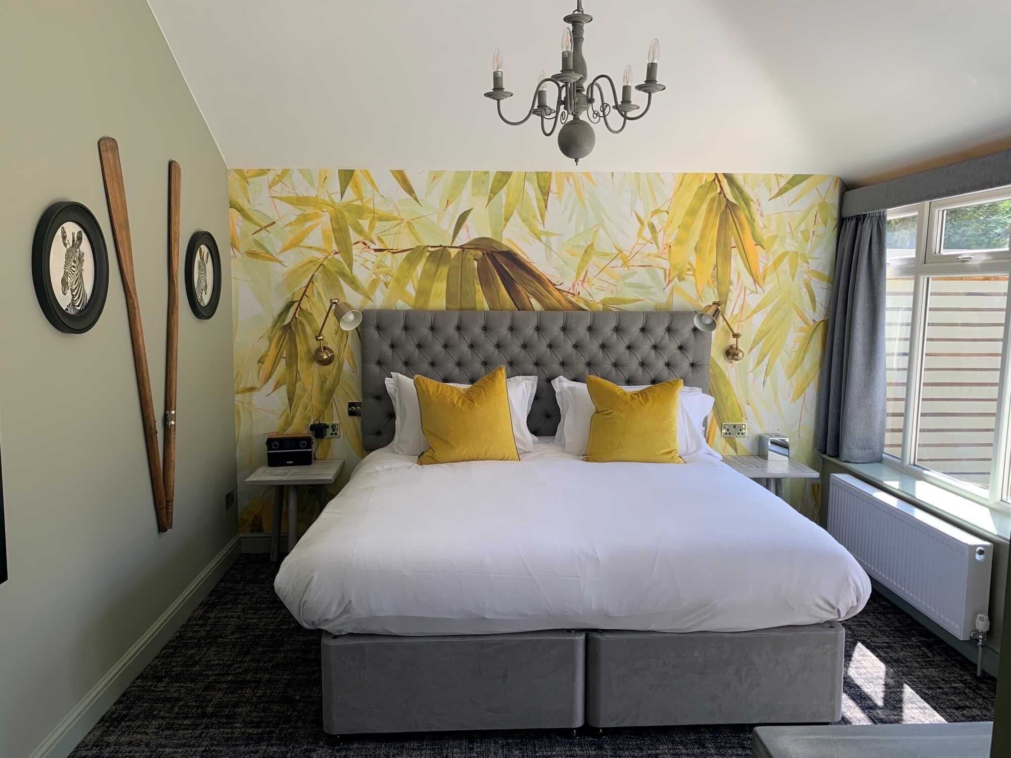Quarto com cabeceira estofada, cama grande com roupa de cama branca e amarela, parede com papel de parede com folhas de bambu, lustre e janela com cortinas.