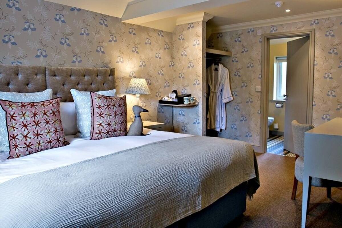 A hotel room with a large bed, a bedside table with a lamp, a chair, and a desk. There is a robe hanging on the door and a small table with a tray of items.
