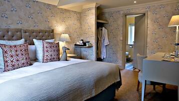 A hotel room with a large bed, a bedside table with a lamp, a chair, and a desk. There is a robe hanging on the door and a small table with a tray of items.