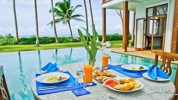 Una mesa de comedor junto a una piscina, con platos de comida, bebidas y un jarrón de flores.