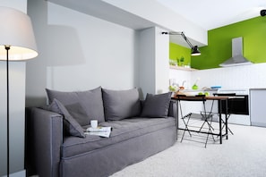 A modern living room with a grey sofa, a small table, and a kitchen area with a stove and green wall. - Live in Athens, Psyrri Central Apartments (Athens)