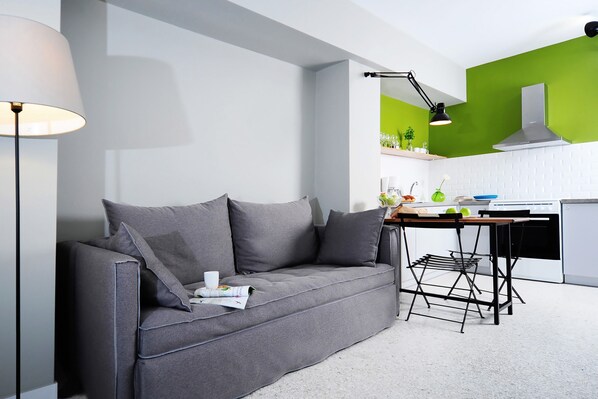 A modern living room with a grey sofa, a small table, and a kitchen area with a stove and green wall. - Live in Athens, Psyrri Central Apartments (Athens)