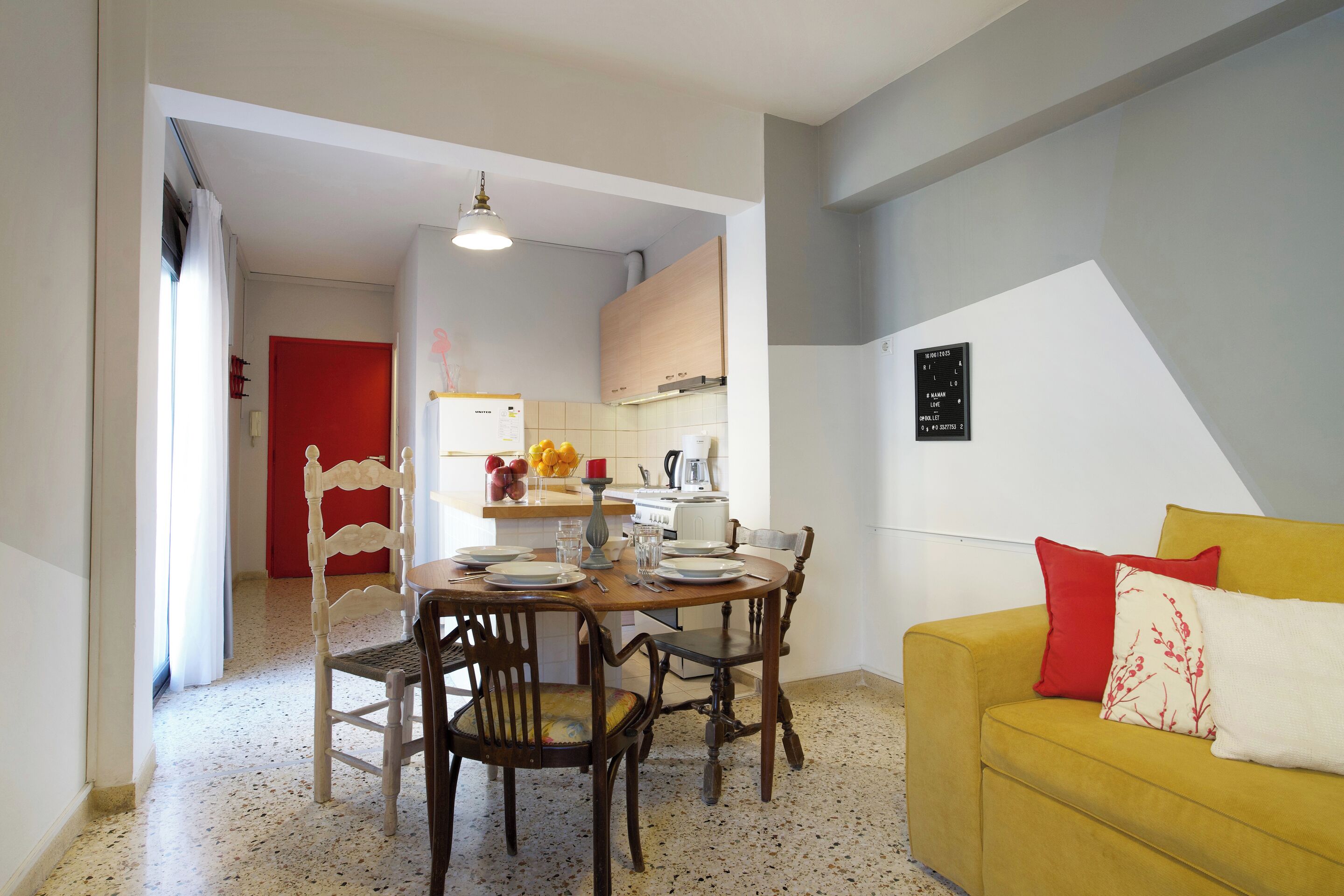 A dining area with a wooden table and chairs, a yellow sofa with red and white cushions, and a kitchen in the background.
