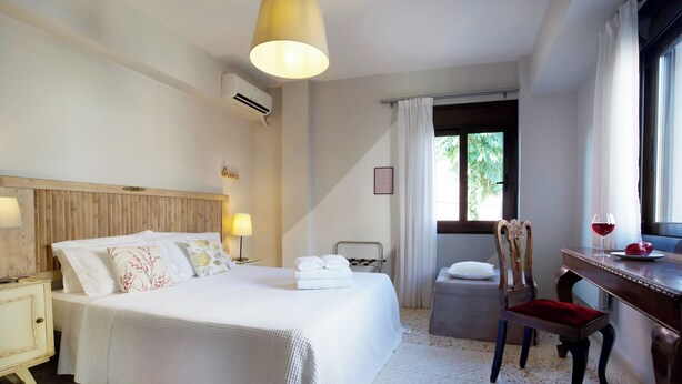 A hotel room with a large bed, a desk with a chair, a window with curtains, and a small table with a wine glass. - Live in Athens, Psyrri Central Apartments (Athens)