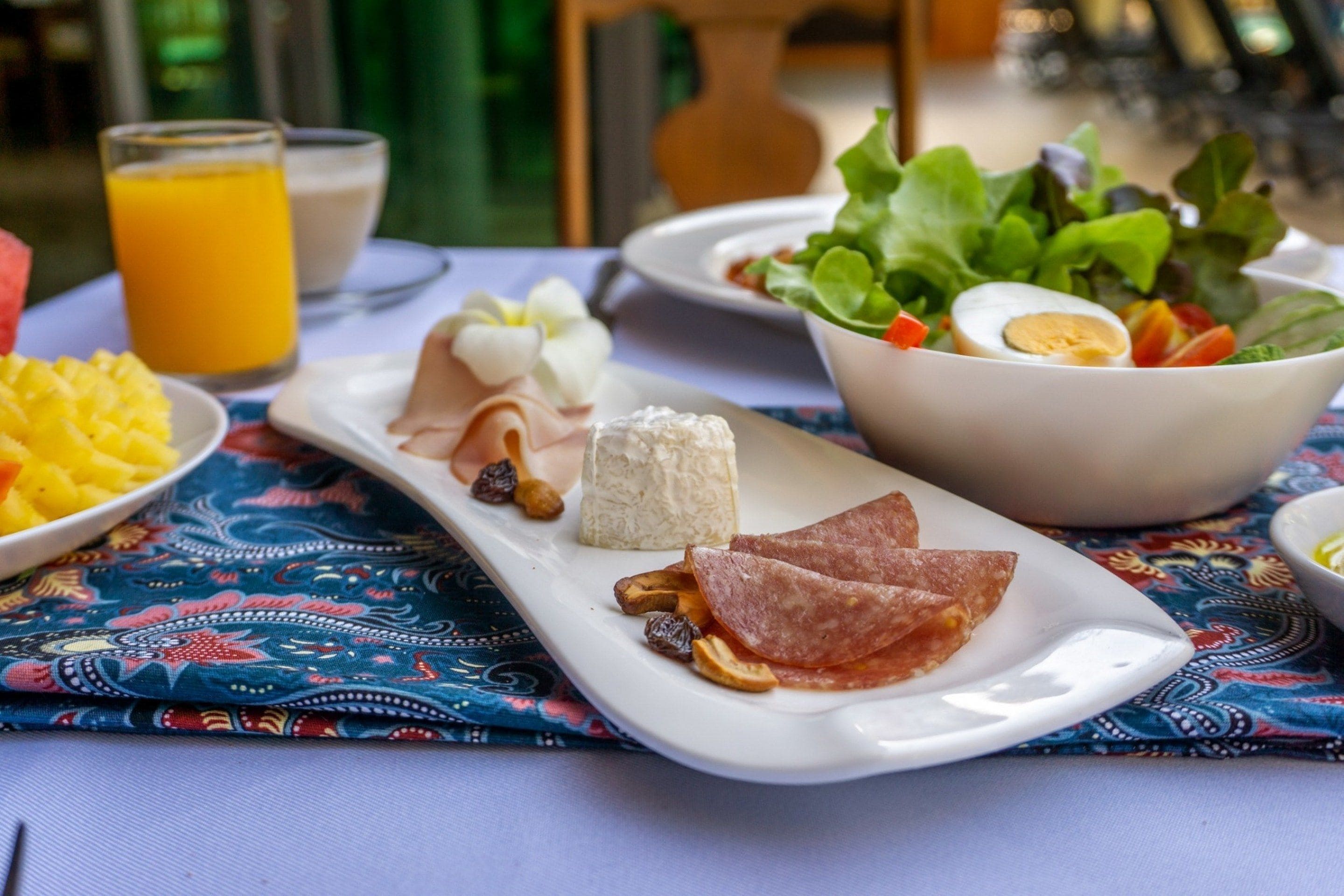 Un desayuno con un plato de jamón, queso y huevos, un bol de ensalada y un vaso de zumo de naranja.