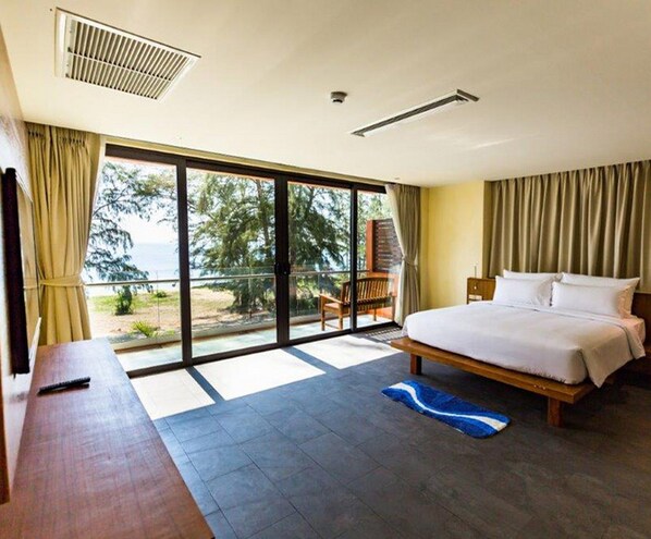 A modern hotel room with a large bed, a desk, and a view of the ocean through floor-to-ceiling windows.
