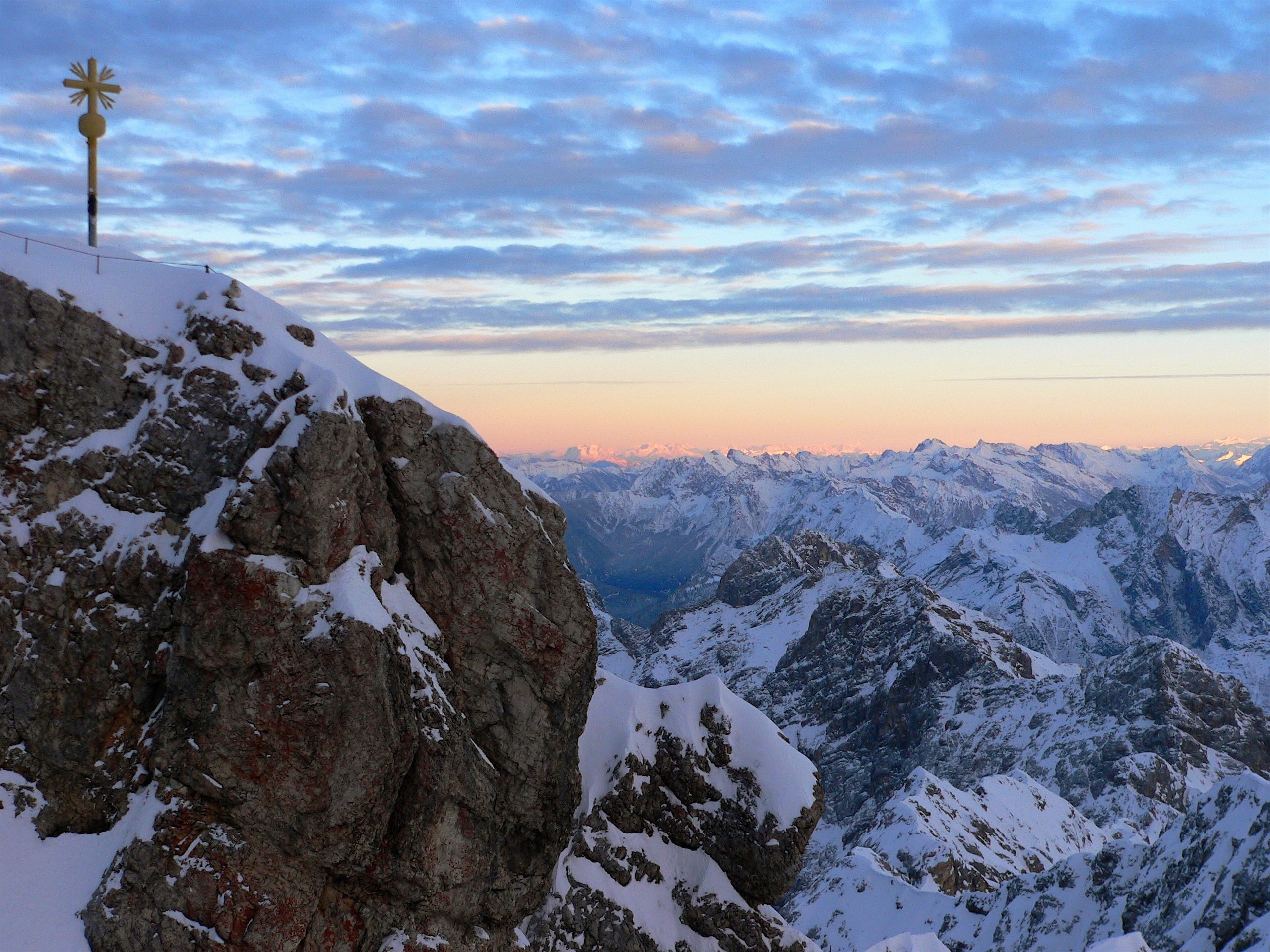 Ein mit Schnee bedeckter Berggipfel mit einem Kreuz auf der Spitze, umgeben von zerklüftetem Gelände und fernen Bergketten unter einem teilweise bewölkten Himmel.