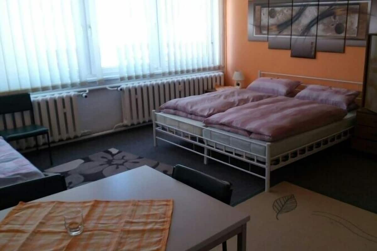 A bedroom with a bed, a desk, a chair, a radiator, and a window with blinds.