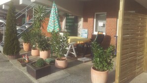 A covered outdoor seating area with potted plants, a table with a yellow tablecloth, and a chair.