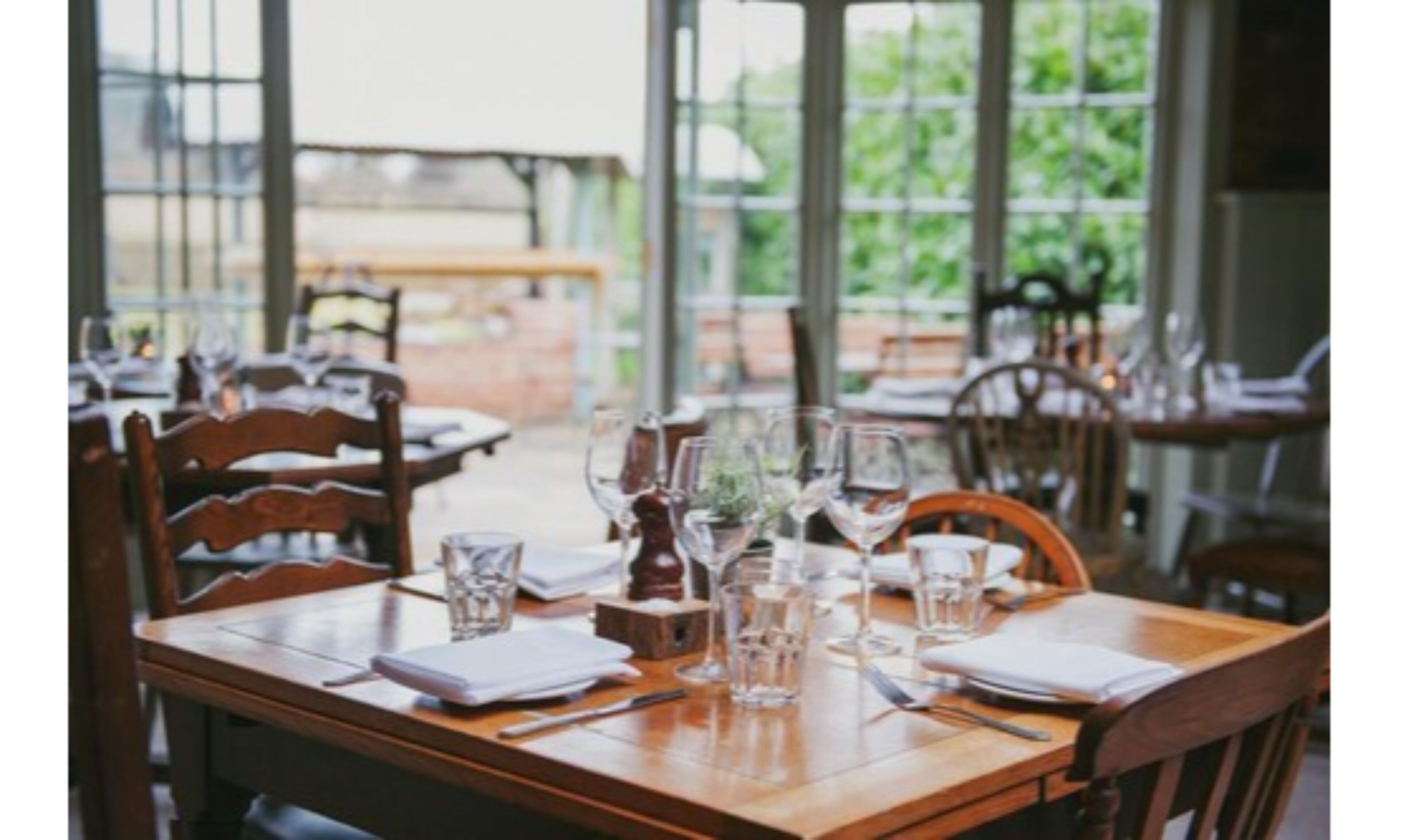 A dining table set with chairs, glasses, and cutlery, with large windows in the background.