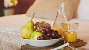 A neatly made bed with a white comforter and pillows, a bowl of fruit, a glass of orange juice, and a bottle of water on a wooden tray.
