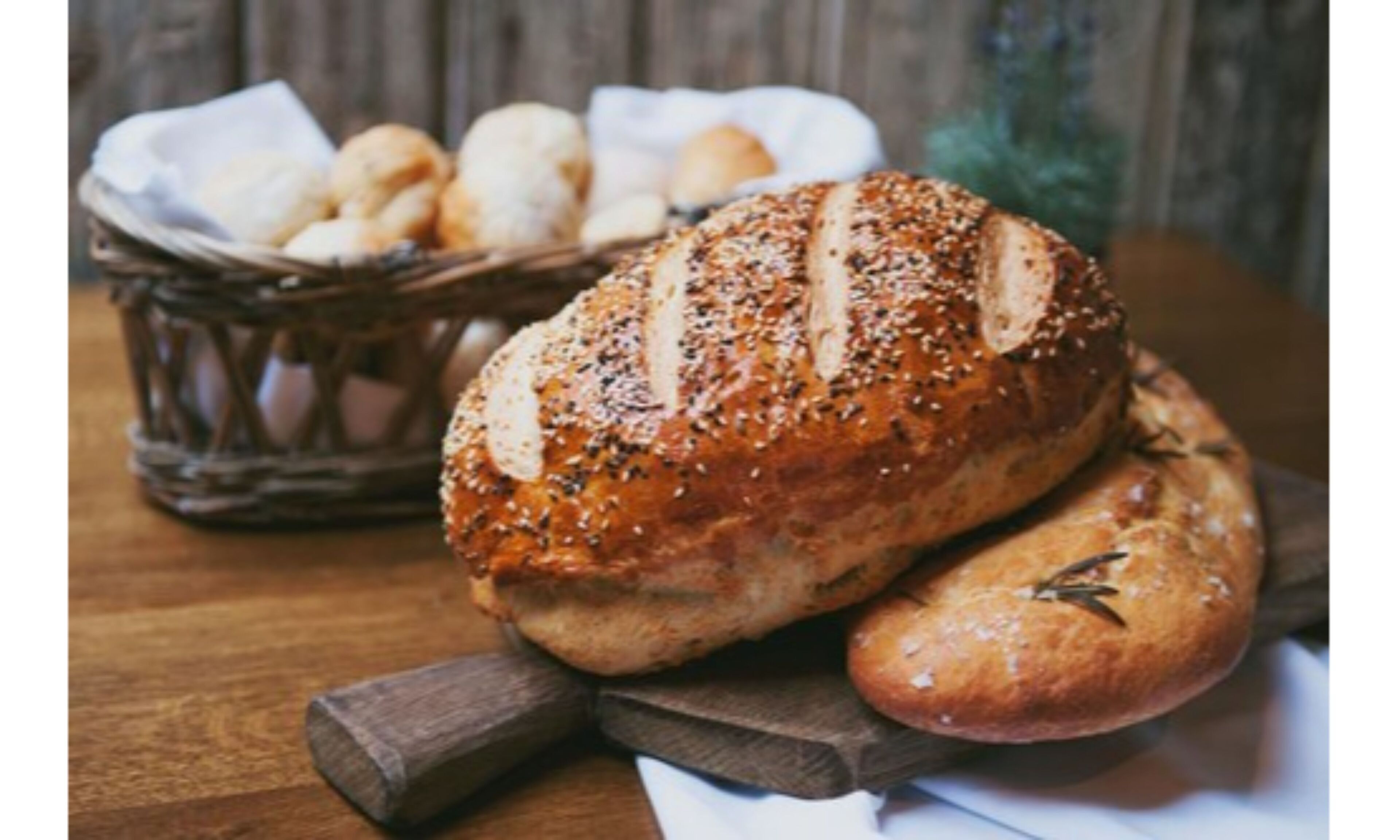 Una hogaza de pan con semillas de sésamo y una cesta de bollos sobre una mesa de madera.