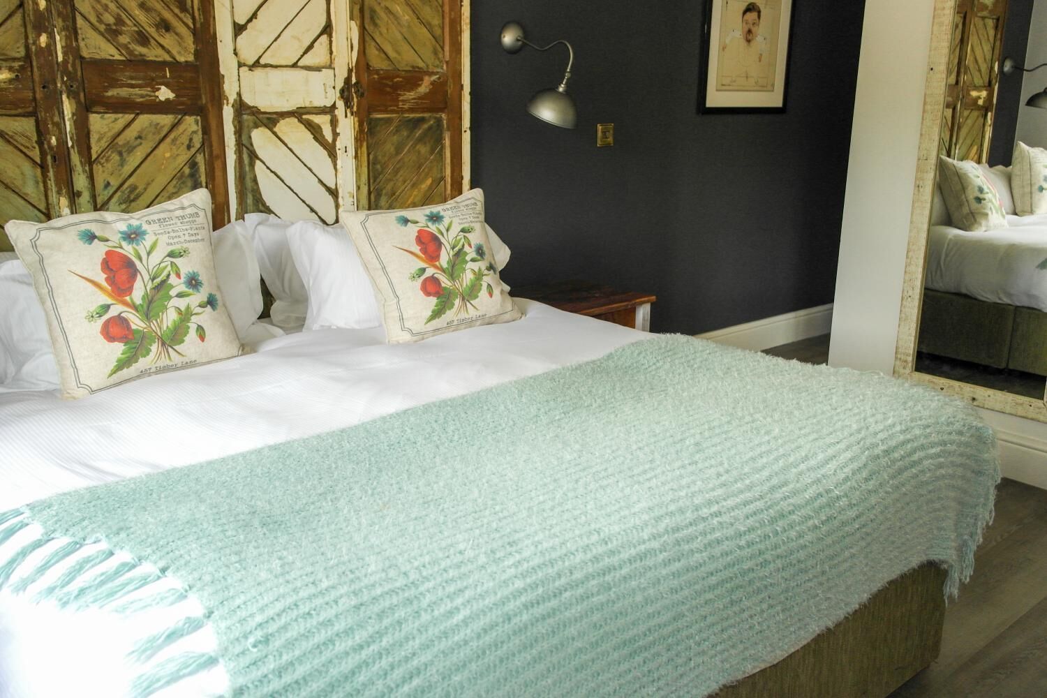 A bed with a white duvet, two pillows with floral designs, a light blue knitted throw, and a wooden headboard.