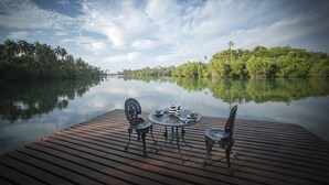Una terraza de madera con una mesa y sillas da a un lago tranquilo, rodeado de abundante vegetación.
