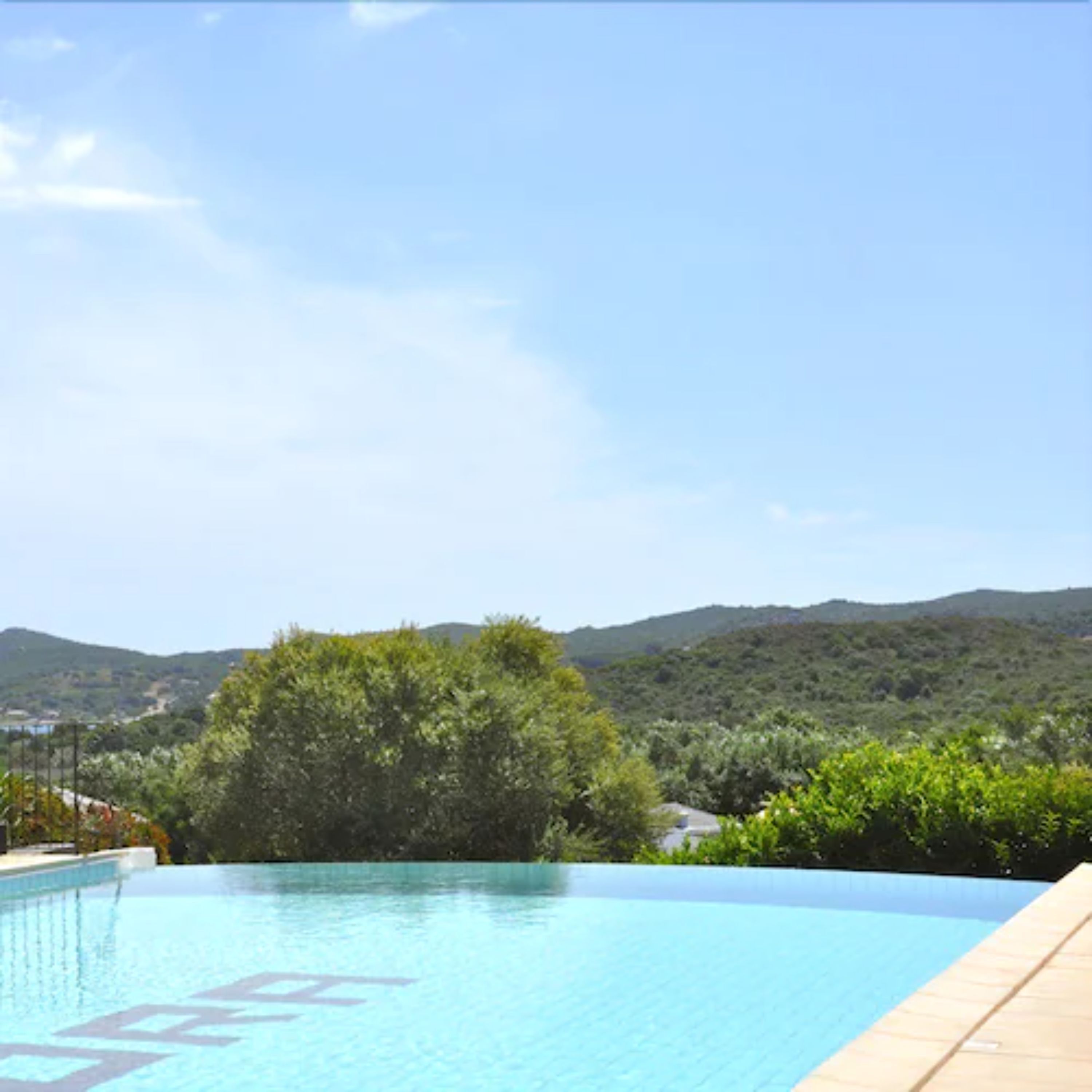 Una piscina azul cristalina con un deck de madera, rodeada de exuberante follaje y colinas al fondo.