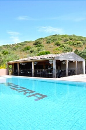A swimming pool with a covered seating area and a hill in the background. - Hôtel Maora Village (Bonifacio)