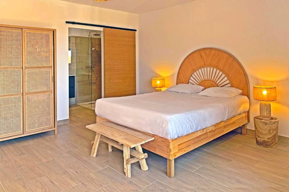 A bedroom with a wooden bed, two bedside lamps, a bench, a rattan wardrobe, and a glass shower door.