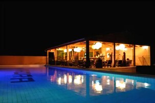 Una zona junto a la piscina con cerramiento de vidrio, mesas, sillas y luces colgantes.