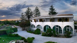 Une propriété située sur la côte, avec un bâtiment blanc doté de fenêtres en arc, un jardin bien entretenu et une vue sur la mer.