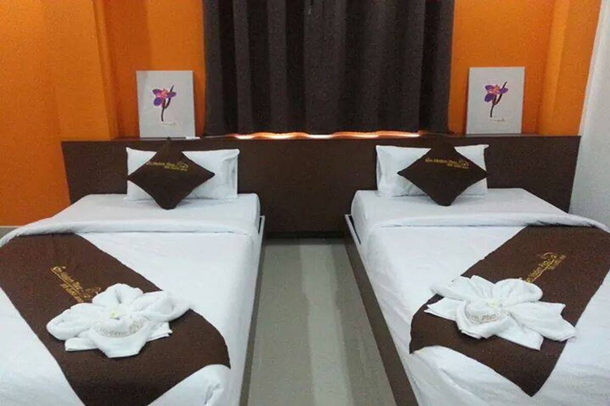 A hotel room with two beds, each with a white bedspread and a brown headboard. There are decorative pillows and a flower arrangement on each bed. The room has orange walls and a dark curtain in the background.