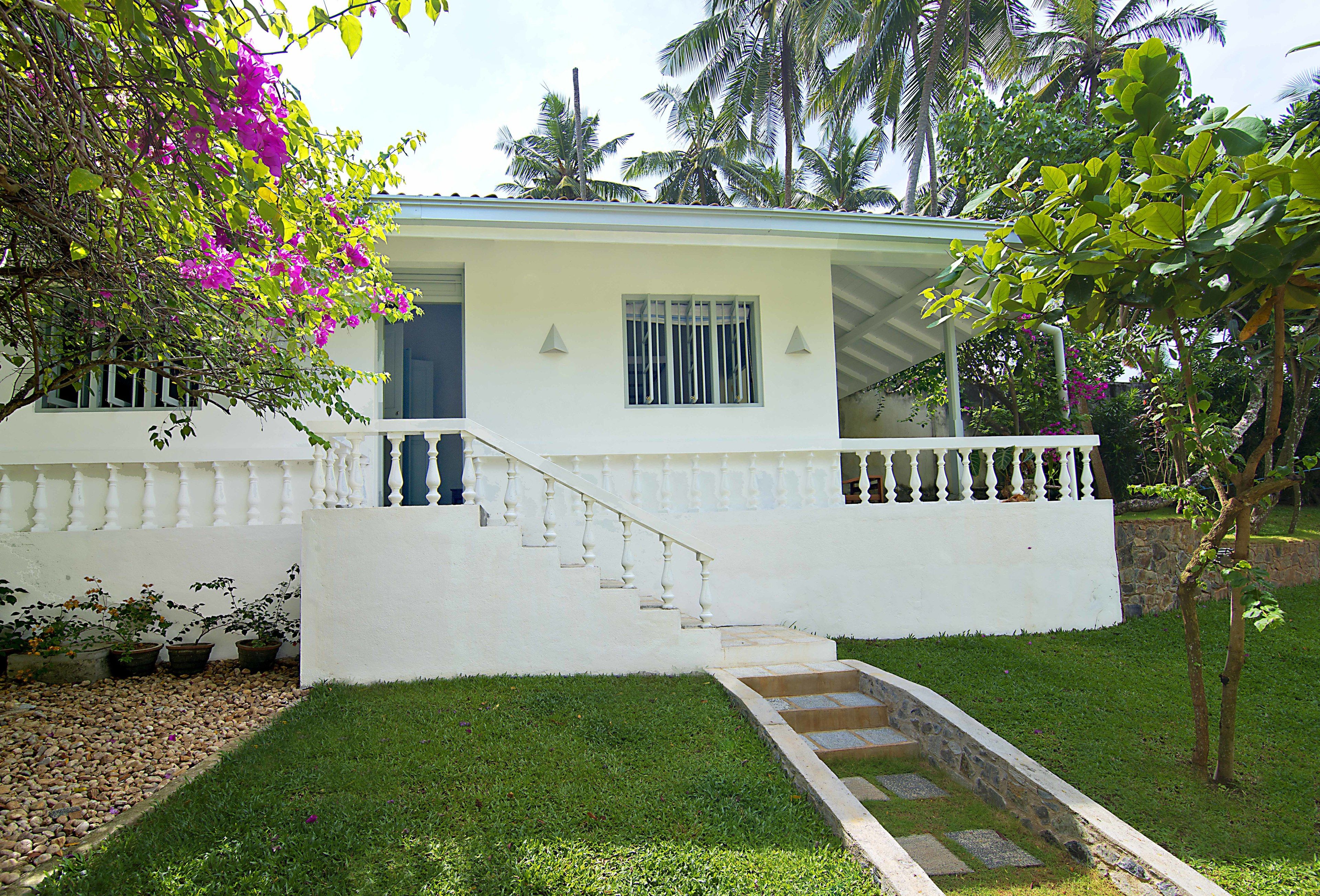 A white house with a front porch, a staircase leading to the entrance, and a small garden area with potted plants and a tree with pink flowers.