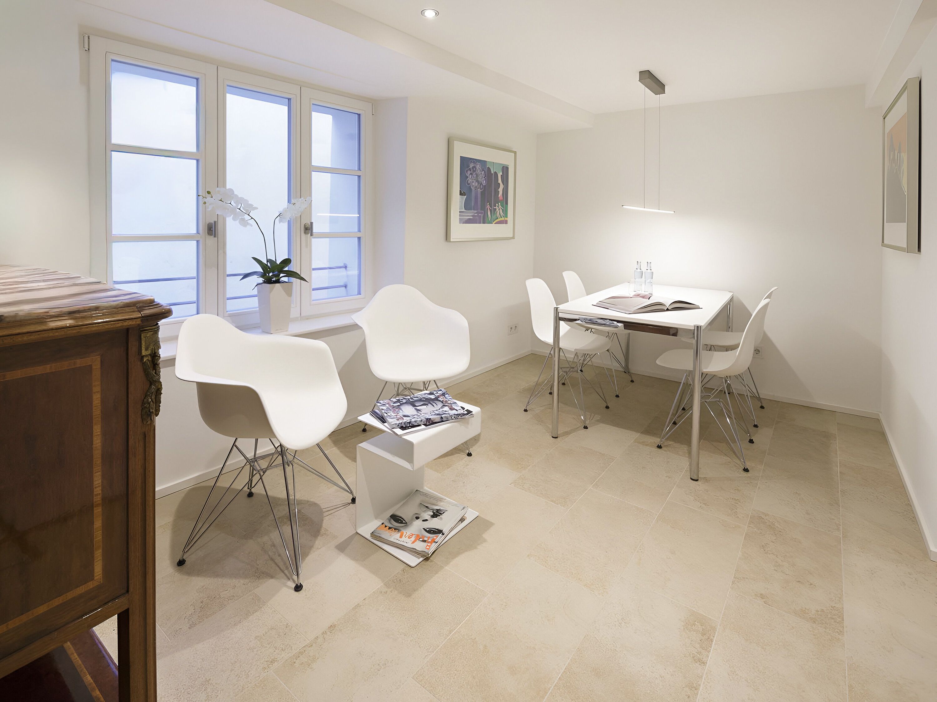 A modern dining area with a white table, four white chairs, a wooden sideboard, and a potted plant by the window.