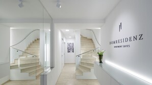 Un moderno vestíbulo de hotel con una escalera de cristal, un mostrador de recepción y un cartel en la pared que dice 'DOMRESIDENZ APARTMENT SUITES'.