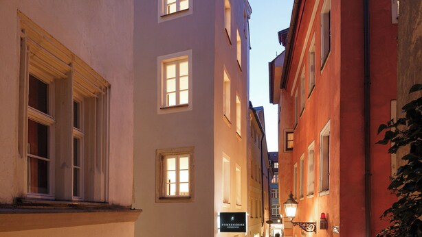 Una estrecha calle adoquinada flanqueada por edificios, incluyendo un hotel llamado "DOMRESIDENZ" y una tienda con un letrero que dice "INSAM".