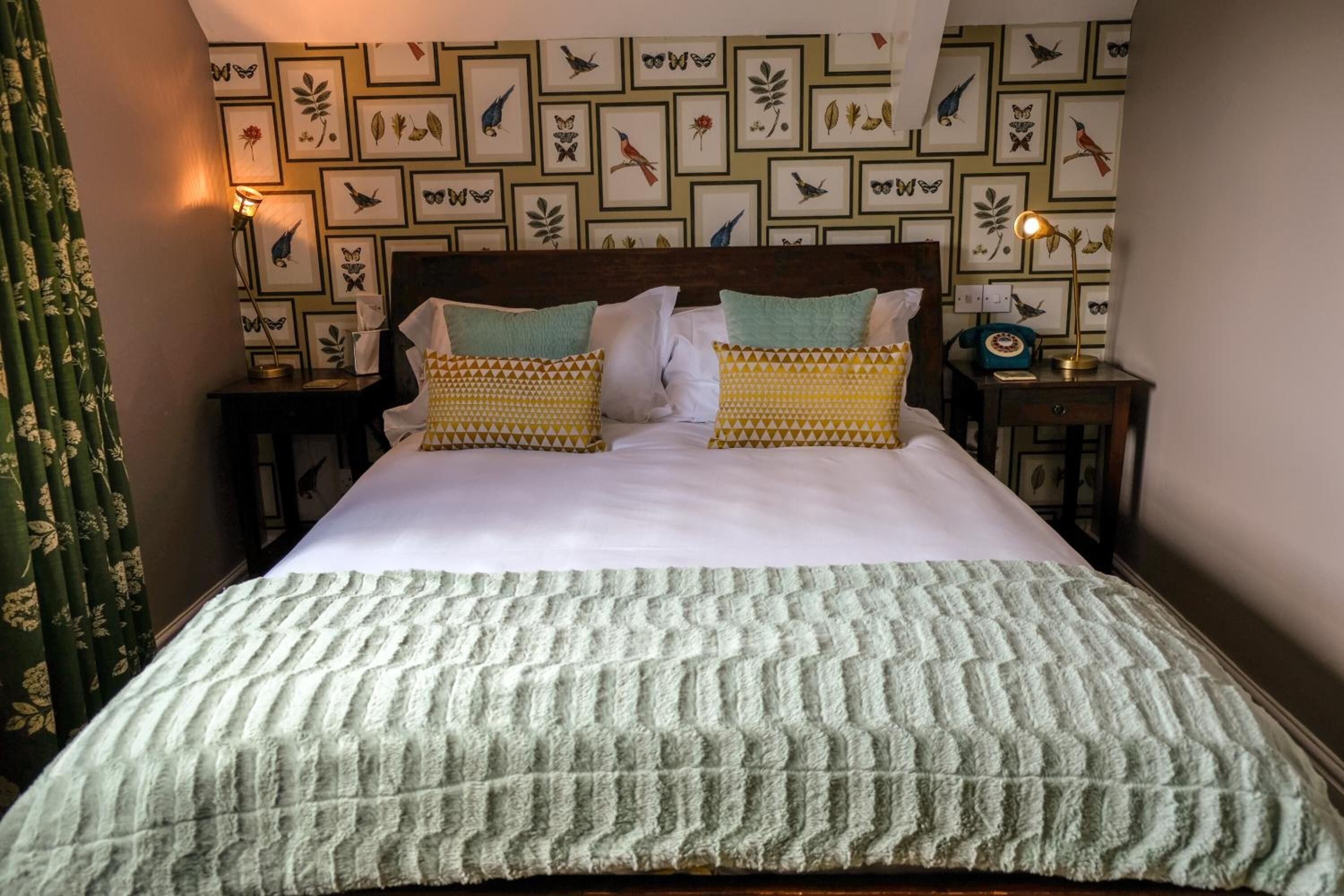 Una cama con cabecera estampada, dos mesitas de noche con lámparas, y una pared decorada con ilustraciones enmarcadas de pájaros y plantas.