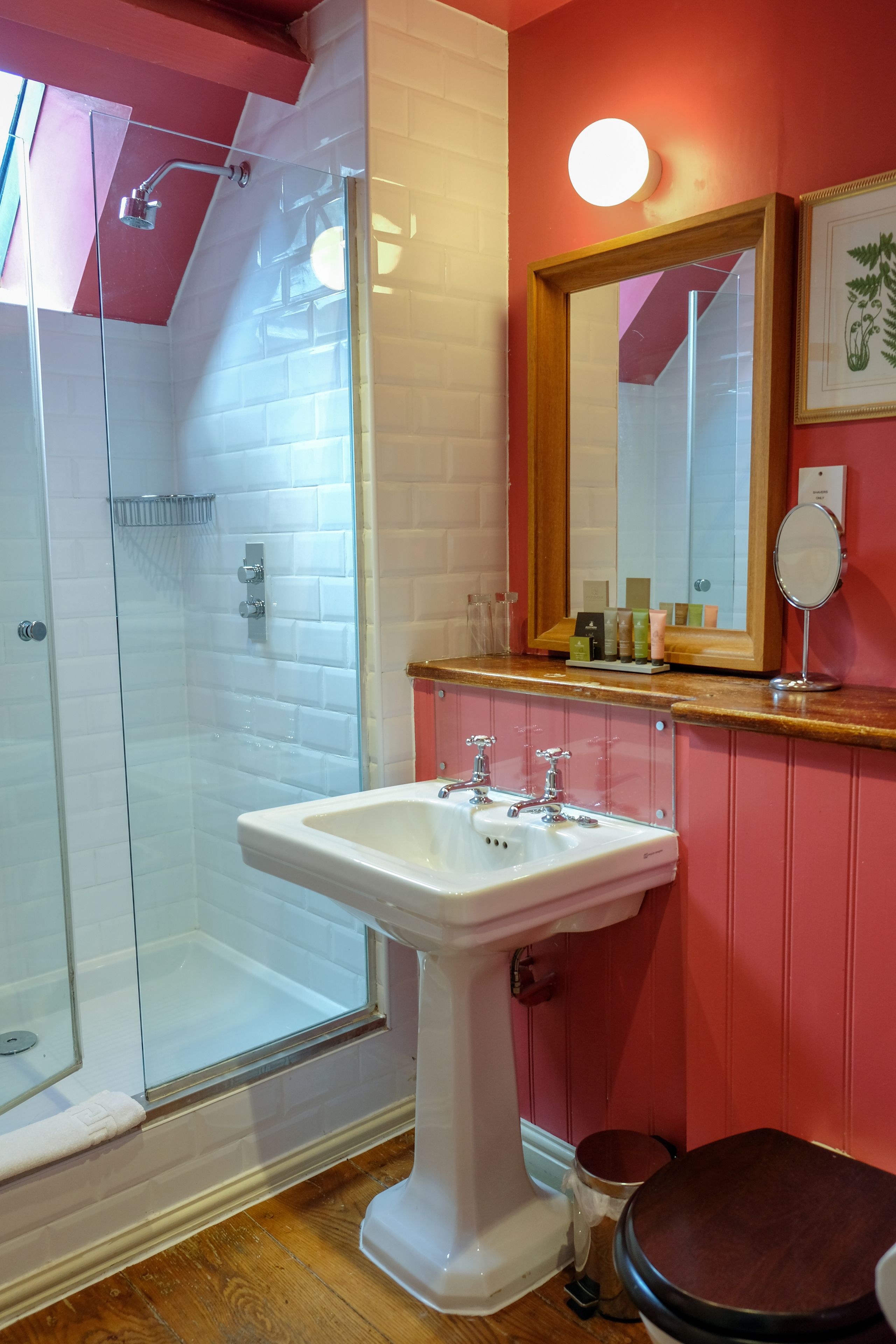 A bathroom with a glass-enclosed shower, a white pedestal sink, a wooden vanity, and a mirror above the sink.