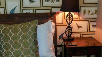 Una cama bien hecha con almohadones estampados, una mesita de noche con lámpara y una pared decorada con ilustraciones enmarcadas de plantas y aves.